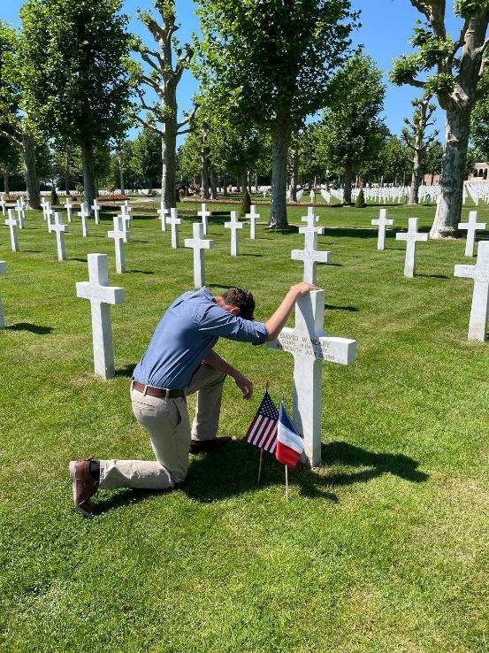 Caleb Deines kneeling at the headstone of his great-uncle, Cpl. David W. Neary, decorated with American and French flags